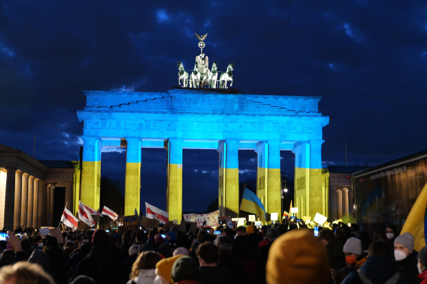 Eine Menschenmenge steht vor dem Brandenburger Tor in Berlin, Deutschland, mit Fahnen und Plakaten, einem Banner auf der rechten Seite und dem Tor mit Statuen und Säulen unter einem bewölkten Himmel.