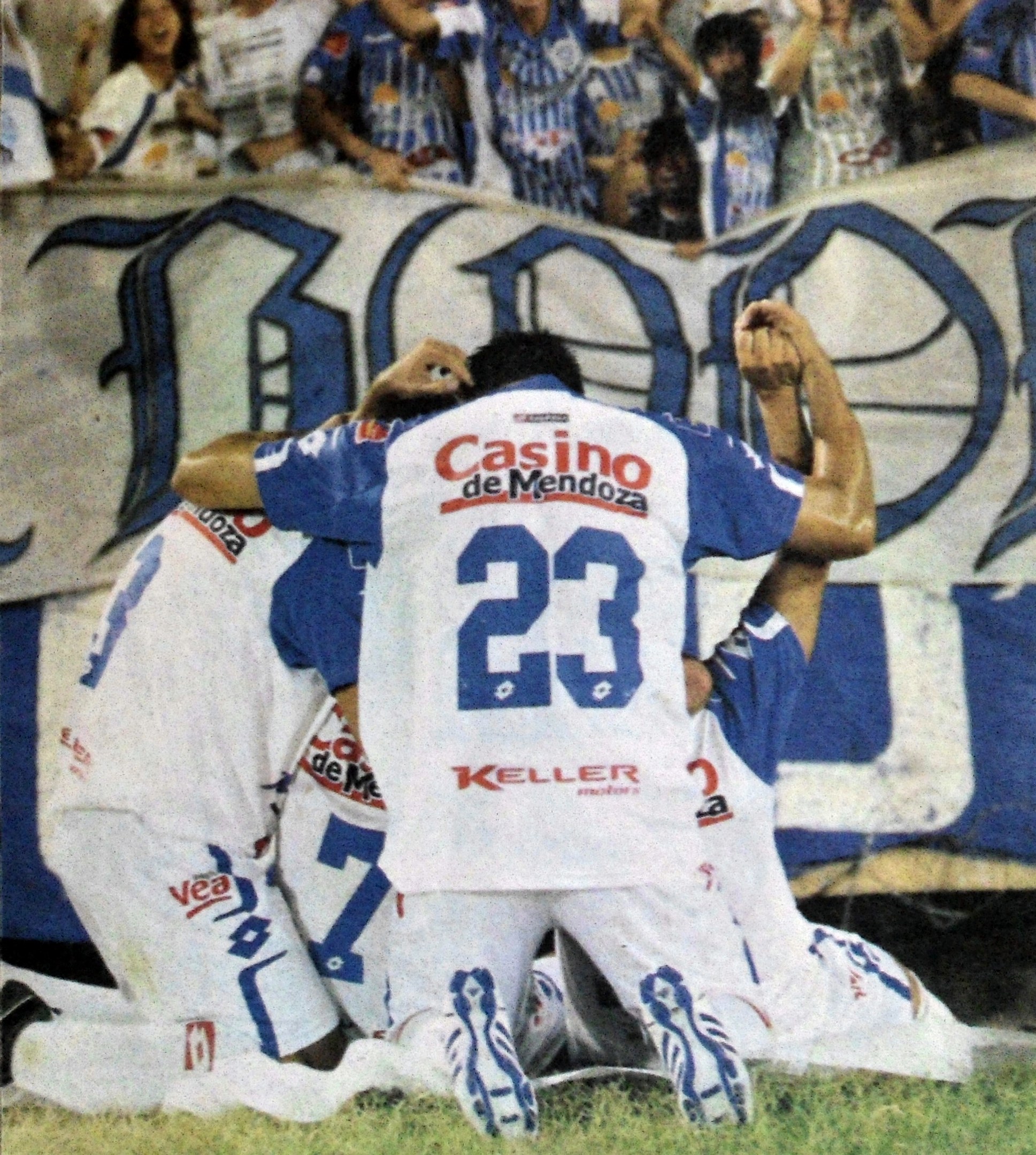 Gruppe von Fußballspielern, die auf dem Boden liegen und mit einer jubelnden Menge feiern, im Hintergrund ist eine teilweise sichtbare Fahne.