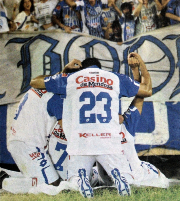 Gruppe von Fußballspielern, die auf dem Boden liegen und mit einer jubelnden Menge feiern, im Hintergrund ist eine teilweise sichtbare Fahne.