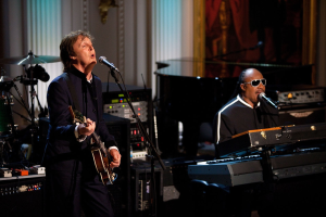 Paul McCartney spielt Gitarre und singt in ein Mikrofon, während Stevie Wonder eine Brille trägt und am Piano spielt, auf der Bühne bei den Grammys, mit Instrumenten und einem gerahmten Bild im Hintergrund.