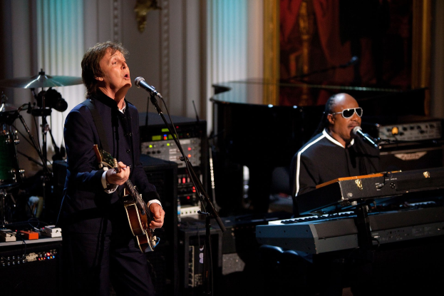 Paul McCartney spielt Gitarre und singt in ein Mikrofon, während Stevie Wonder eine Brille trägt und am Piano spielt, auf der Bühne bei den Grammys, mit Instrumenten und einem gerahmten Bild im Hintergrund.