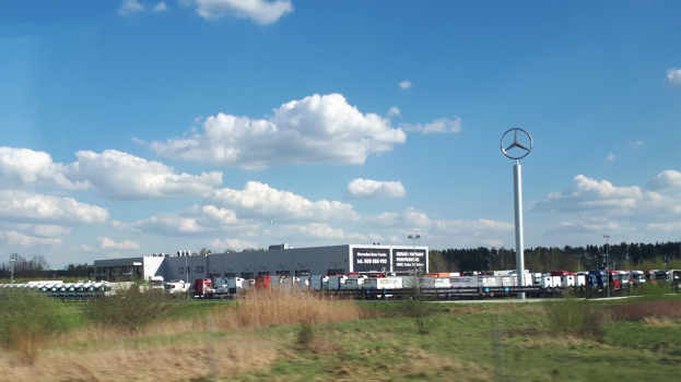 Mercedes-Benz-Fabrik in Deutschland mit Fahrzeugen, Grünflächen, Gebäuden und einem Logo-Mast unter einem bewölkten Himmel.