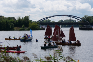 Eine Gruppe von Menschen in kleinen Booten auf einem Gewässer, mit einer Brücke, Fahrzeugen, Bäumen, einem bewölkten Himmel im Hintergrund und Pflanzen unten, die ein Regatta suggerieren.