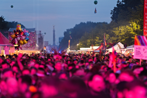 Eine große Menschenmenge auf einem nächtlichen Musikfestival, beleuchtet von bunten Lichtern und Ballons, mit Zelten, Bannern, Fahnen und anderen Gegenständen, die herumliegen, und Bäumen, einem Turm und einem sternenklaren Himmel im Hintergrund.
