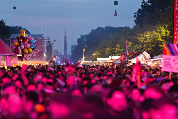 Eine große Menschenmenge auf einem nächtlichen Musikfestival, beleuchtet von bunten Lichtern und Ballons, mit Zelten, Bannern, Fahnen und anderen Gegenständen, die herumliegen, und Bäumen, einem Turm und einem sternenklaren Himmel im Hintergrund.