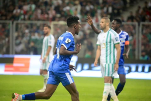 Ein Fußballspieler in einem blauen und weißen Trikot feiert ein Tor, umgeben von einer Gruppe in weißen und grünen Trikots, mit einem unscharfen Hintergrund, der ein weißes und blaues Board und ein Geländer zeigt.