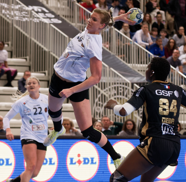 Eine Gruppe von Frauen, die auf einem Platz Handball spielen, mit einem Ball in der Luft, Zuschauern auf Treppen und Geländern und einem "Handball-Weltmeisterschaft 2019 - Tag 2"-Schild im Hintergrund.