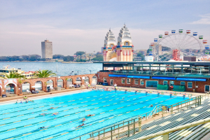 Großer Swimmingpool mit schwimmenden Menschen, umgeben von Geländern und Bänken, mit einem Riesenrad, Gebäuden, Bäumen und einem bewölkten Himmel im Hintergrund.