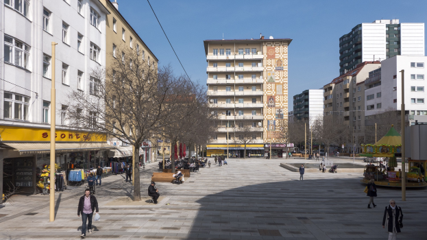 Ein belebter Stadtplatz mit Fußgängern, Bänken, Bäumen, Gebäuden, einem Karussell und Strommasten unter einem sichtbaren Himmel.