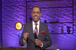 Stephen Colbert in Anzug, Krawatte und Brille auf einer Bühne mit Mikrofon stehend, mit einer Wand und verschiedenen Gegenständen dahinter.