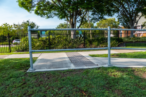 Ein Metallzaun in einer grünen Fläche, umgeben von Pflanzen, Bäumen und einem Auto auf der Straße, mit Gebäuden und einem klaren blauen Himmel im Hintergrund.