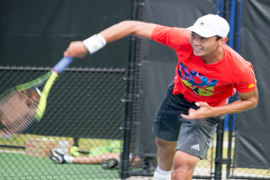 Ein Mann in roter Jacke und weißer Mütze spielt Tennis, hält einen Schläger, mit einem Netz und durchscheinendem Hintergrund; eine weitere Person sitzt in der Nähe auf dem Boden mit einer Box und Bällen.