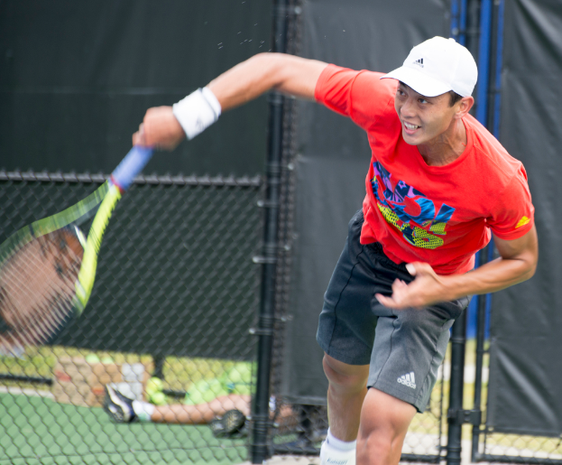 Ein Mann in roter Jacke und weißer Mütze spielt Tennis, hält einen Schläger, mit einem Netz und durchscheinendem Hintergrund; eine weitere Person sitzt in der Nähe auf dem Boden mit einer Box und Bällen.
