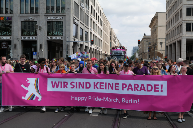 Gruppe von Menschen marschiert auf einer Berliner Straße mit einem pinken "Happy Pride March"-Schild, mit Gebäuden, Laternenmasten und Verkehrszeichen an der Straße unter einem bewölkten Himmel.