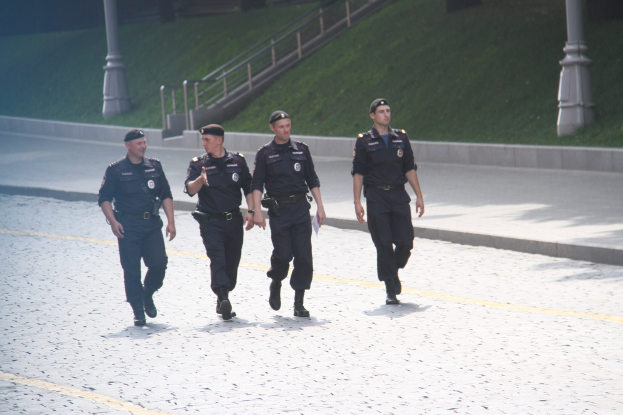 Drei Polizisten in Uniform und mit Mützen gehen eine Straße entlang mit Polen, Geländern, Treppen und Gras im Hintergrund.