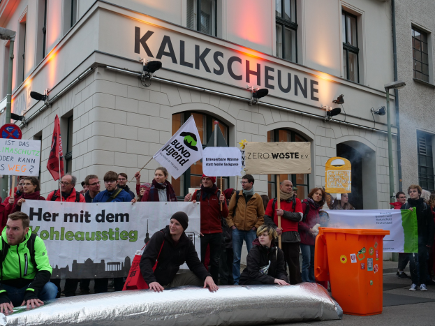 Eine Gruppe von Menschen mit Protestschildern und -plakaten vor einem Gebäude, mit zwei Personen im Vordergrund und einem Müllcontainer auf der rechten Seite.