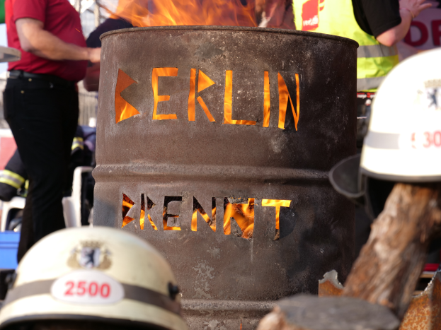 Eine Gruppe von Menschen um ein Feuerhydrant mit der Aufschrift 'Berlin' herumstehend mit einem brennenden Fass im Vordergrund und Helmen unten und einem hölzernen Objekt auf der rechten Seite.