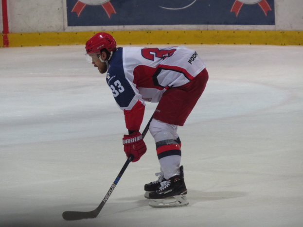 Ein Hockey-Spieler in voller Ausrüstung fährt auf dem Eis Schlittschuh, hält einen Stock und hat ein Banner mit Text und Logos im Hintergrund.