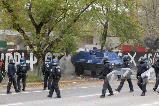 Polizisten in schwarzer Uniform und Helmen gehen neben einem gepanzerten Fahrzeug eine Straße entlang, einige halten Schilde, mit Bäumen, Gebäuden und einem klaren blauen Himmel im Hintergrund.