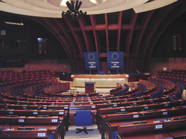 Großer halbrunder Konferenzraum des Europäischen Parlaments in Brüssel mit einem zentralen Podium, Sitzreihen und Tischen sowie Texttafeln an den Wänden unter einer beleuchteten Decke.