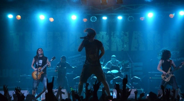 Eine Gruppe von Musikern, die auf einer hell erleuchteten Bühne in der O2 Arena in London spielen, mit einem Sänger, der ein Mikrofon hält, anderen, die Gitarren spielen, und einem Schlagzeuger im Hintergrund, während man unten die Hände des Publikums sieht.