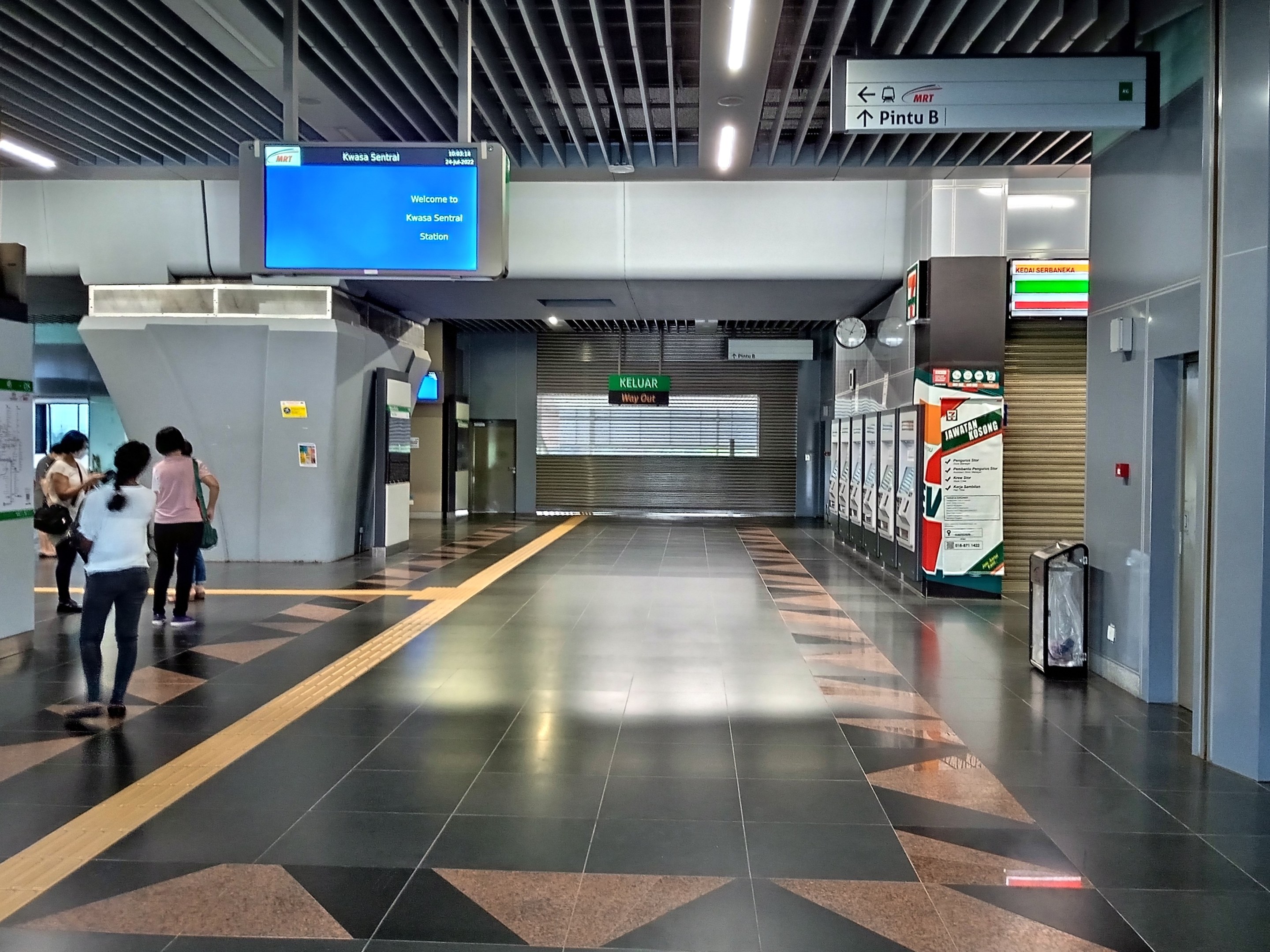 Menschen gehen durch einen Bahnhof mit einem Wagen auf der rechten Seite, Schilder mit Text, eine Uhr an der Wand und Deckenleuchten.