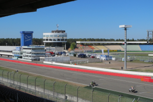 Gruppe von Motorradfahrern auf einer Rennstrecke, umgeben von einem Zaun, Fahrzeugen, einem Gebäude, Schildern, Pfosten, einem Stadion, Beleuchtung, Bäumen und einer bewölkten Himmel.