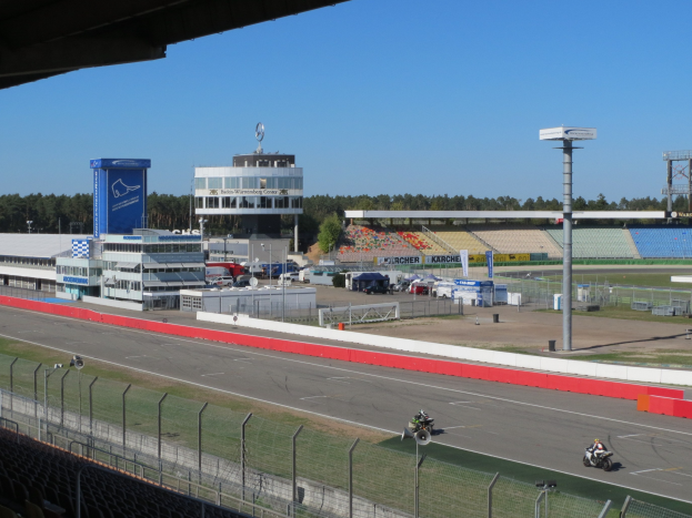 Gruppe von Motorradfahrern auf einer Rennstrecke, umgeben von einem Zaun, Fahrzeugen, einem Gebäude, Schildern, Pfosten, einem Stadion, Beleuchtung, Bäumen und einer bewölkten Himmel.