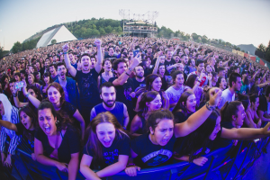 Eine große Menschenmenge auf einem Musikfestival hebt die Hände vor einem Geländer, mit Bäumen, Hütten und Lichtern im Hintergrund unter einem klaren blauen Himmel, einschließlich eines Mannes und einer Frau.