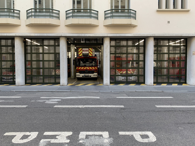 Feuerwache mit Feuerwehrauto vor der Tür, mit Fenstern, Geländern, Säulen, Glastüren und einer Straße unten.