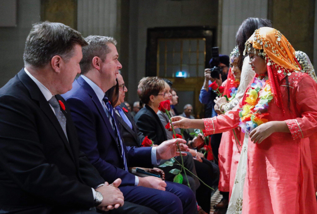 Eine Gruppe von Menschen sitzt in einem Raum zusammen, mit einer Frau in der Mitte, die einen Kranz trägt und Blumen hält, flankiert von einem Mann im Anzug und jemandem, der eine Kamera hält, vor einer Wand mit einer Tür.