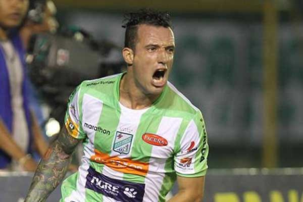 Ein Fussballer in grün-weißer Uniform mit Tattoos auf dem Arm feiert nach einem Tor, mit unscharfen Hintergrundfiguren und einer Tafel.