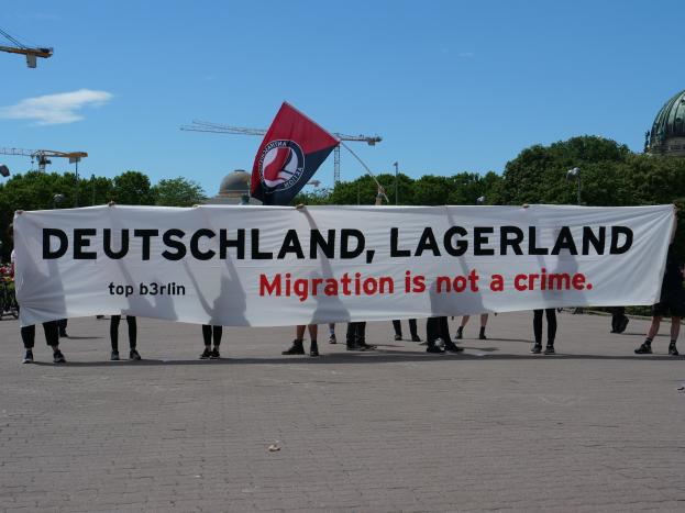Eine Gruppe von Menschen mit einem Transparent "Deutschland, Lagerland Migration ist kein Verbrechen" und einer Flagge, im Hintergrund Straßenpfähle, Straßenlaternen, Bäume, Kräne, ein Gebäude und ein bewölktes Himmel.