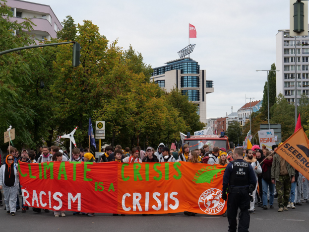 Eine Gruppe von Menschen marschiert auf einer von Bäumen gesäumten Straße, die eine Tafel mit der Aufschrift "Klimakrise ist eine Krise" trägt, mit parkenden Fahrzeugen, Schildern und Gebäuden im Hintergrund unter einem klaren blauen Himmel.