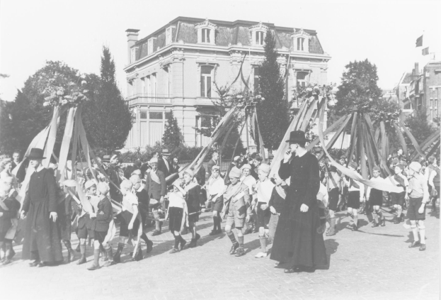 Schwarzes und weißes Bild einer Gruppe von Menschen, die eine Straße entlanggehen, einige halten Fahnen, mit Hüten, Bäumen, Gebäuden und einem sichtbaren Himmel im Hintergrund, das einen Maypole zeigt, der auf ein May Day-Fest hinweist.