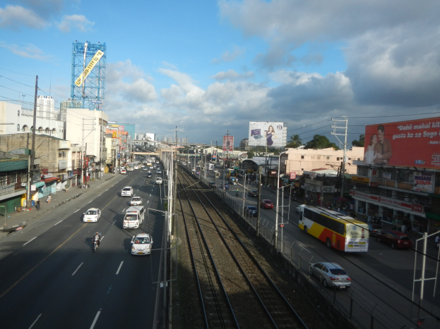 Stadtstraße mit Schwerlastverkehr neben einer Bahnschiene, mit Fahrzeugen, Strommästen, Gebäuden, Schildern, Bäumen und einem bewölktem Himmel.