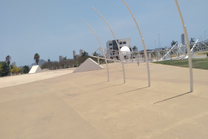 Ein Skaterpark mit einer zentralen Metallstruktur, umgeben von Pfosten, Gras, einem Zaun, Bäumen, Gebäuden, Straßenmasten und einem bewölktem Himmel.