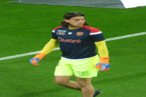 Ein Mann in einem blauen Hemd und gelben Shorts läuft auf einem Fußballfeld und streckt die Arme aus.