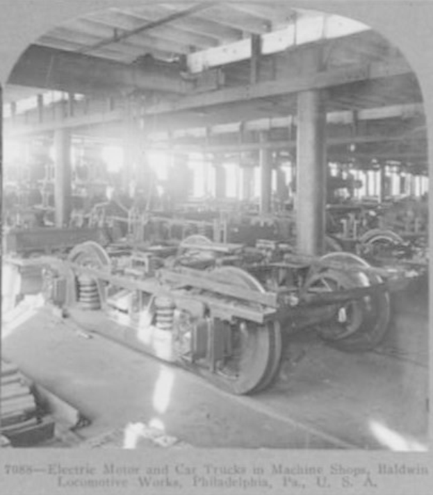 Ein Schwarz-Weiß-Foto eines Elektromotors und eines Lastwagens in einer Fabrik, die von Maschinen, Säulen und einem Dach erfüllt ist, mit Text am unteren Rand.