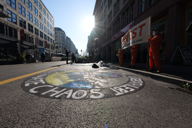 Eine Gruppe von Menschen mit Protestschildern und Plakaten auf einer Stadtstraße mit Gebäuden, Pflanzen und einer gemalten "Chaos"-Nachricht, die auf Occupy Wall Street verweist.
