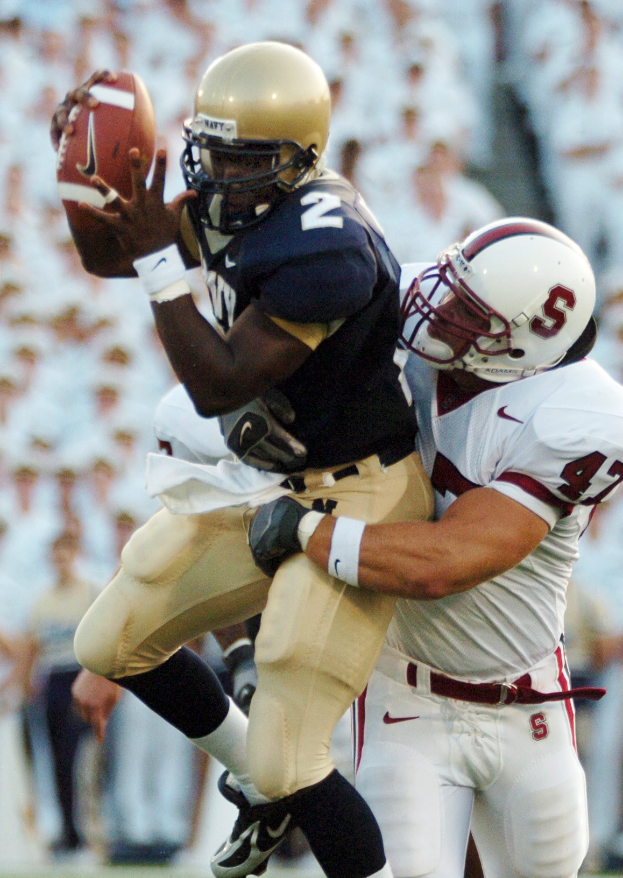 Zwei Fütballspieler in Helmen und Uniformen: einer fängt einen Ball in der Luft, würend er von einem anderen getackelt wird, mit einem unscharfen Hintergrund.