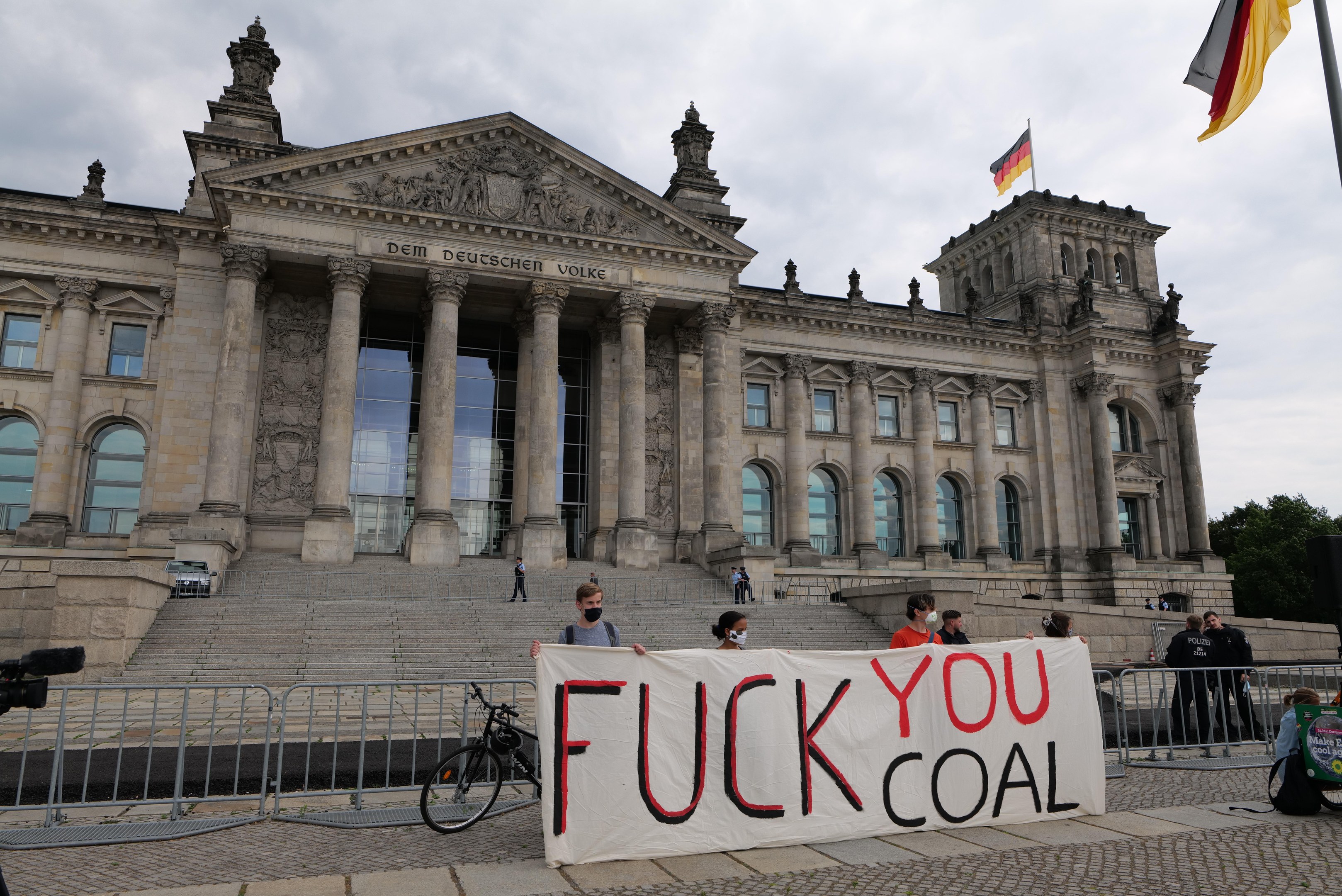 Protestierende halten ein "Fuck You Coal"-Schild vor dem Reichstaggebäude in Berlin, Deutschland, mit den architektonischen Details des Gebäudes, einem Fahrrad in der Nähe, Bäumen, einem Fahnenmast und einem bewölkten Himmel.