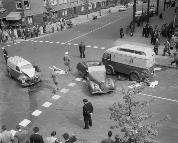 Schwarzes und weißes Bild eines Autounfalls an einer Straßenkreuzung mit mehreren Fahrzeugen, darunter ein Van, und einer Gruppe von Menschen, die daneben stehen, mit Gebäuden, Bäumen, Pfählen und Texttafeln im Hintergrund.