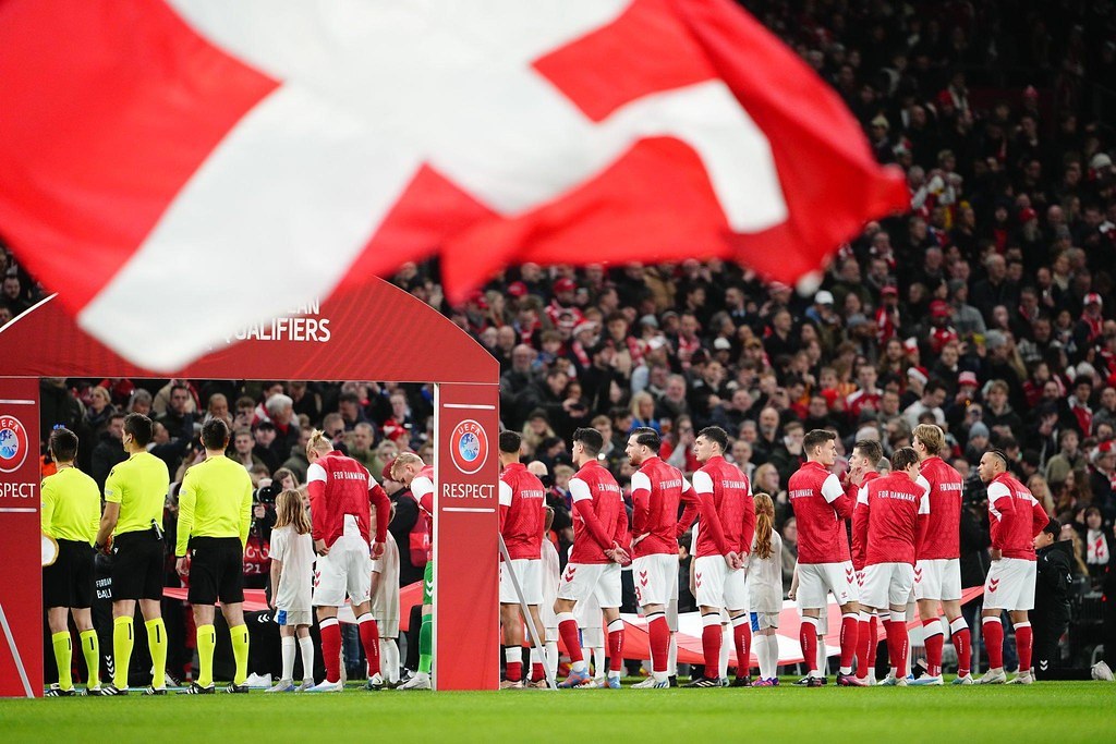Gruppe von Menschen auf einem Fußballfeld mit einer roten und weißen Flagge im Vordergrund, einem Bogen mit dem Text "Bayern München vs. Bayern München Wetten & Vorschau" im Hintergrund und einer großen Menge im Stadion.
