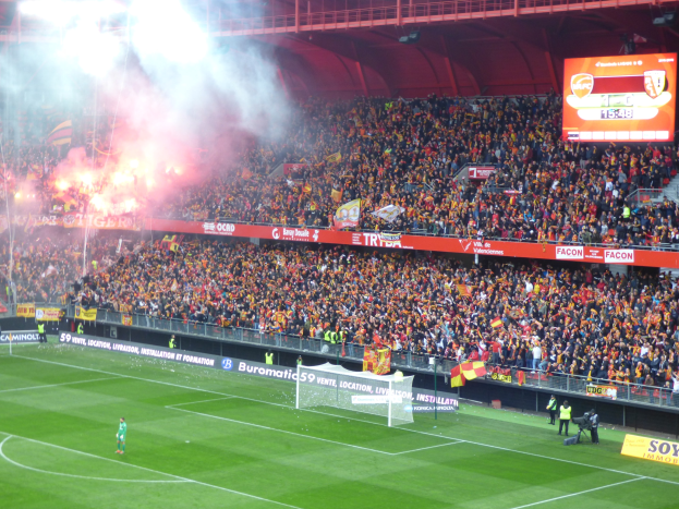 Ein Fußballspiel in einem großen Stadion mit Spielern auf dem Boden in der Nähe eines Torpfostens, umgeben von Zäunen, Bannern, Fahnen, einem Anzeigebildschirm, Metallrahmen, einem Dach mit Lampen und aufsteigendem Rauch.