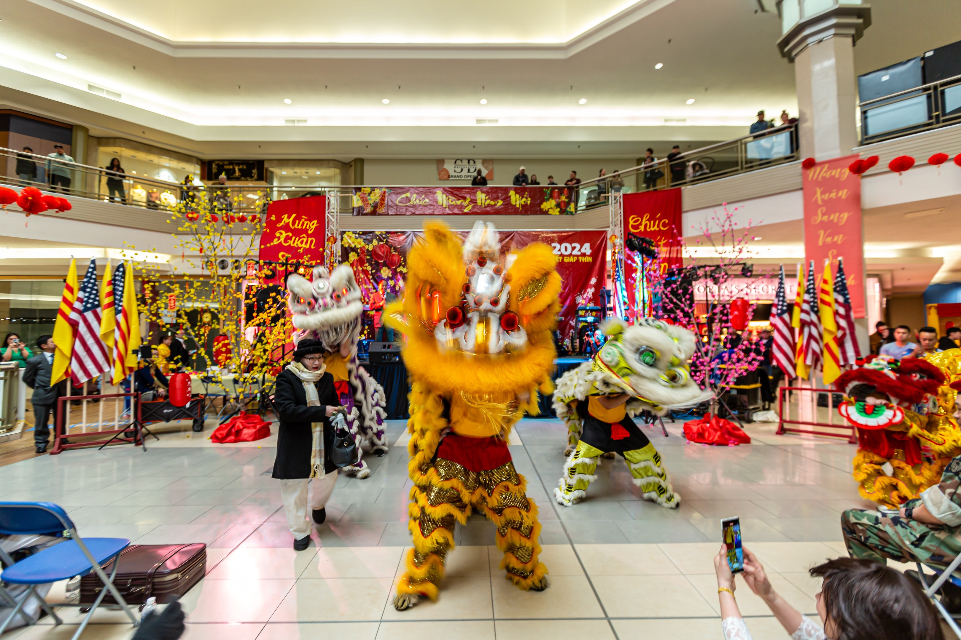 Ein lebendiges chinesisches Neujahrsfest in einem Einkaufszentrum mit tanzenden Menschen in Kostümen, anderen auf Stühlen sitzend und Handys nutzend, umgeben von Fahnen, Bannern, Dekorationen und Deckenlampen.
