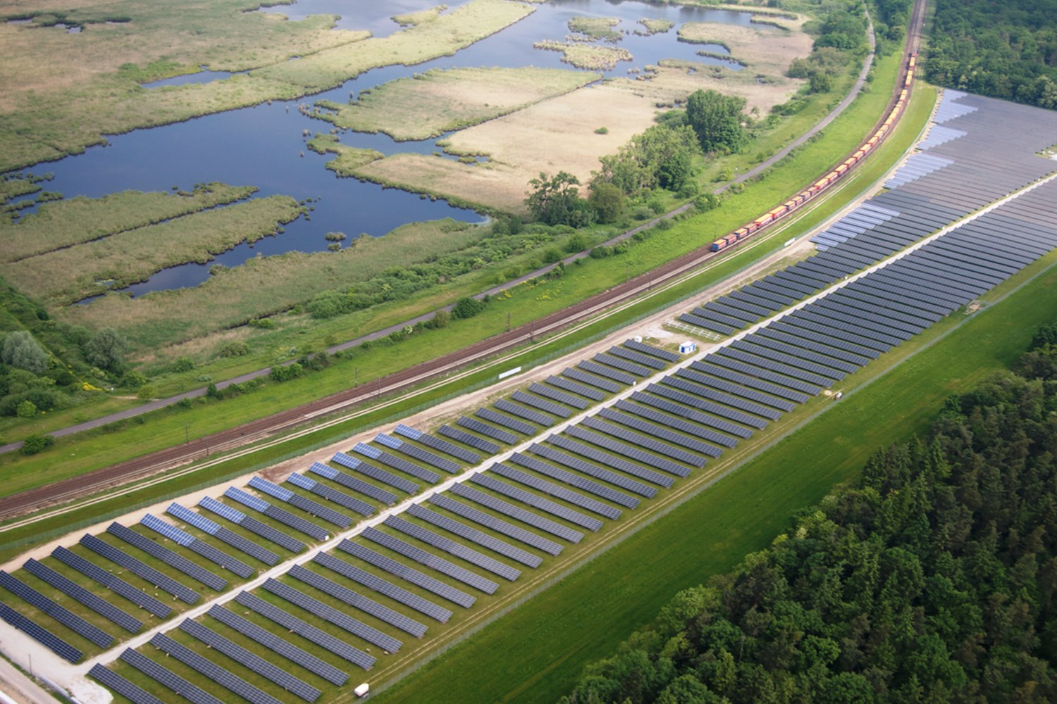 Luftaufnahme einer Solar-Farm mit Panels, Bäumen, Gras und Wasser, mit einem Zug auf einer nahen Bahnstrecke.