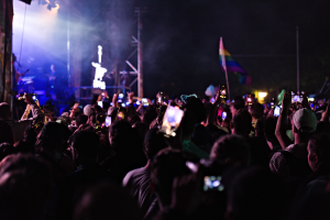 Menschenmenge auf einem Konzert mit hochgehaltenen Handys, beleuchtet von Scheinwerfern, umgeben von Pfosten und einer Fahne, mit Musikern auf der Bühne.