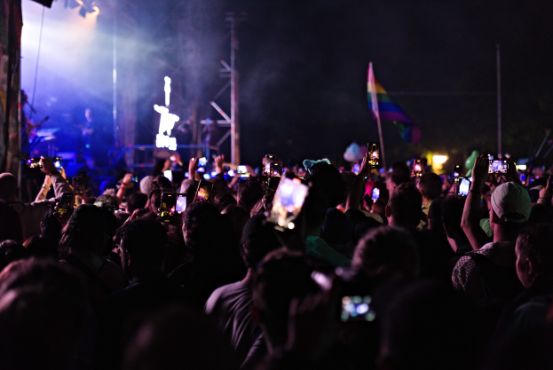 Menschenmenge auf einem Konzert mit hochgehaltenen Handys, beleuchtet von Scheinwerfern, umgeben von Pfosten und einer Fahne, mit Musikern auf der Bühne.