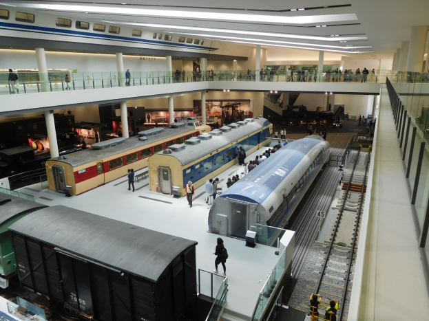 Eine geräumige Museumsausstellung zeigt verschiedene Züge in unterschiedlichen Größen und Farben, bei der Besucher vom Boden, Geländern und Säulen unter Deckenlampen beobachten.